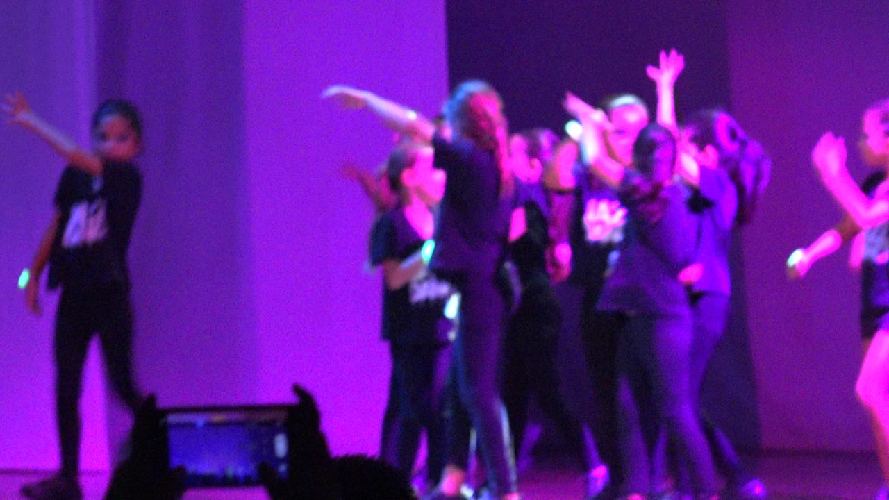 Dance group raises arms together under bright purple stage lighting.
