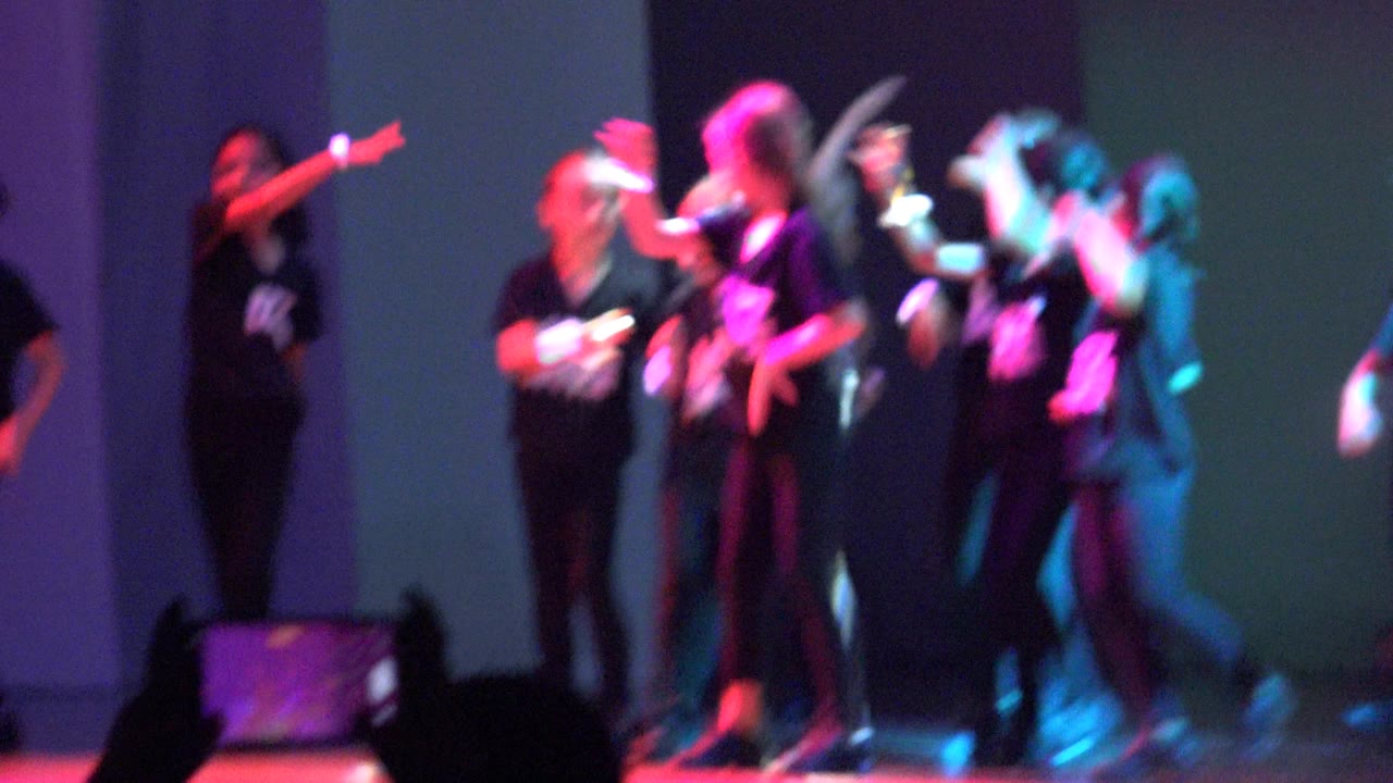 Young performers in black Jazz Dance shirts cluster on a stage in San Jose Del Cabo BCS Mexico, while one dancer gestures sideways and teal light hits the right side of the group.