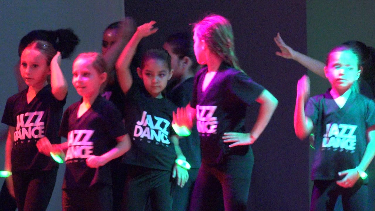 Children perform a jazz dance routine in San Jose Del Cabo, Mexico, with several raised arms, black shirts, and pink and green stage lighting.