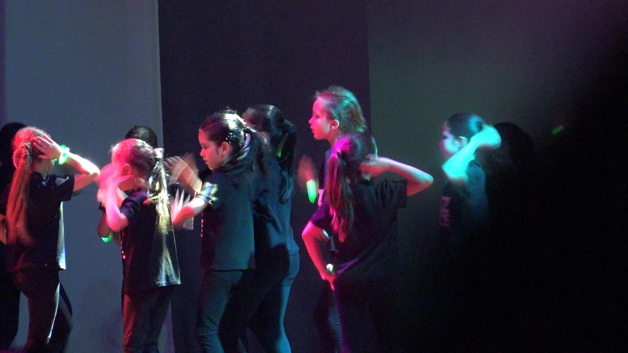 Children in matching Jazz Dance shirts face each other on the indoor stage in San Jose del Cabo BCS Mexico, with green and pink highlights catching their hands and hair.