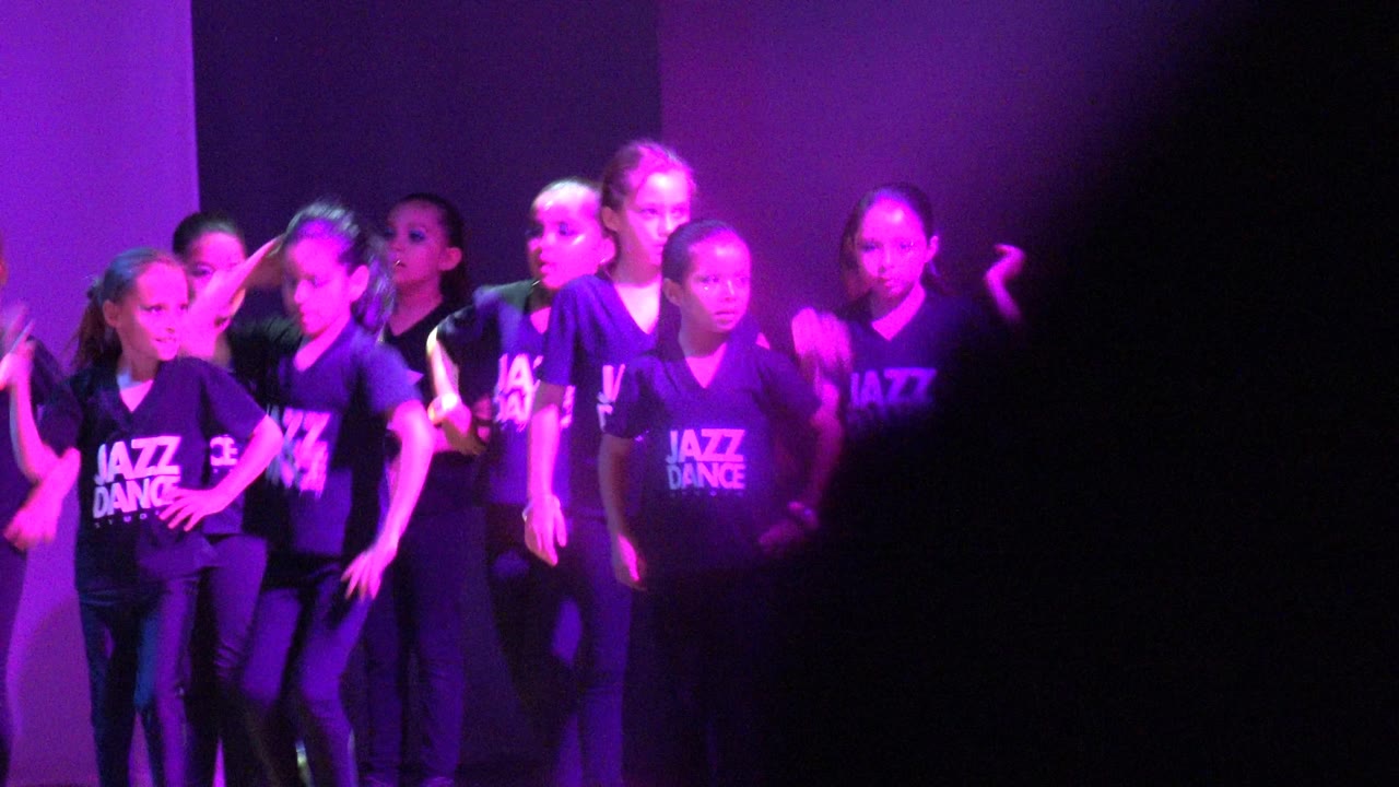 Children performing a jazz dance routine with arms raised under purple lighting.