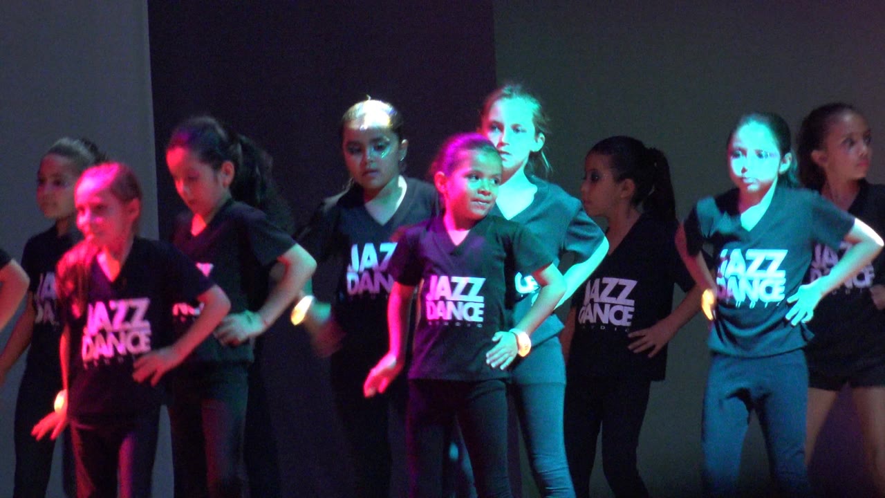 Children in black Jazz Dance shirts stand across a stage in San Jose Del Cabo BCS Mexico, with several hands on hips under pink and cyan light.