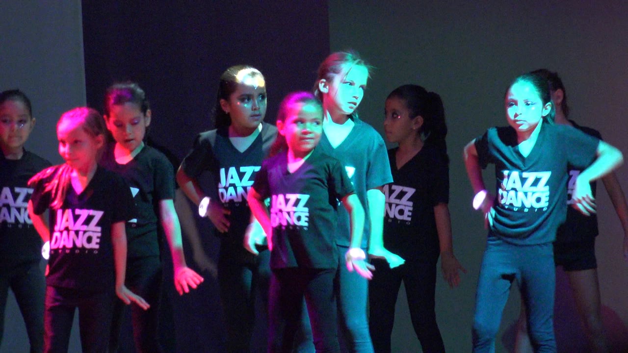 Children in Jazz Dance shirts face forward on a stage in San Jose Del Cabo BCS Mexico, with one front dancer lit cyan and red while others hold hip and arm positions.