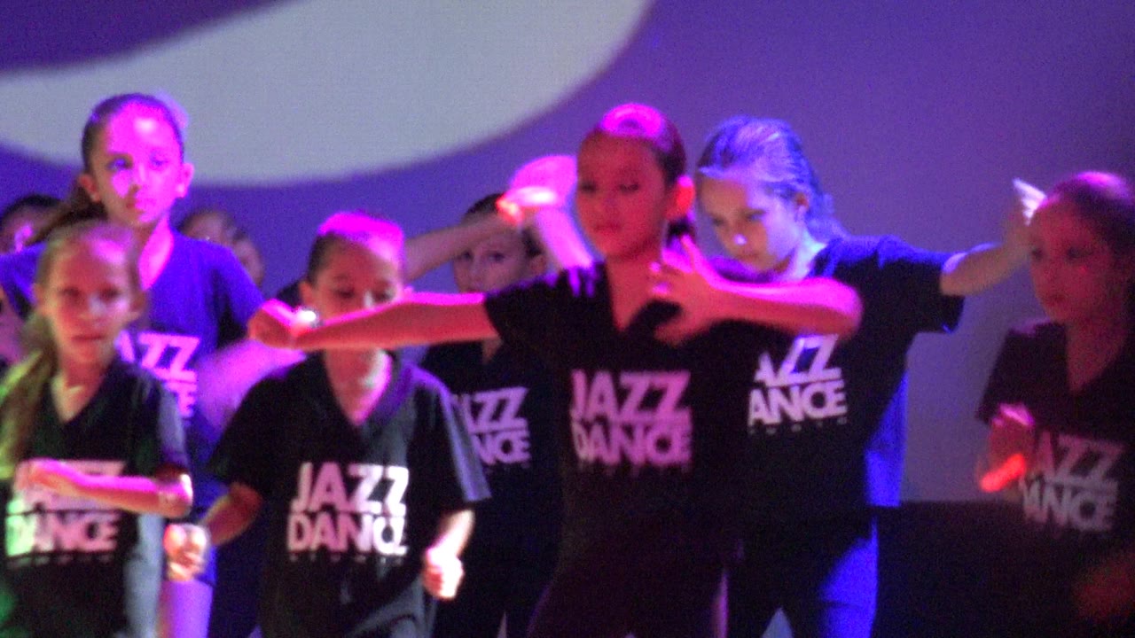 Children in Jazz Dance shirts extend their arms during the routine in San Jose del Cabo, Mexico, with glow sticks catching purple, blue, and pink stage light.