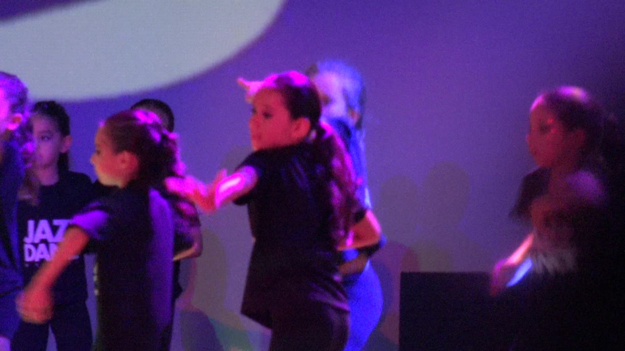 Children perform a jazz dance routine on a purple-lit stage in San Jose del Cabo, Mexico, with black shirts and several visible faces near the left side of the frame.