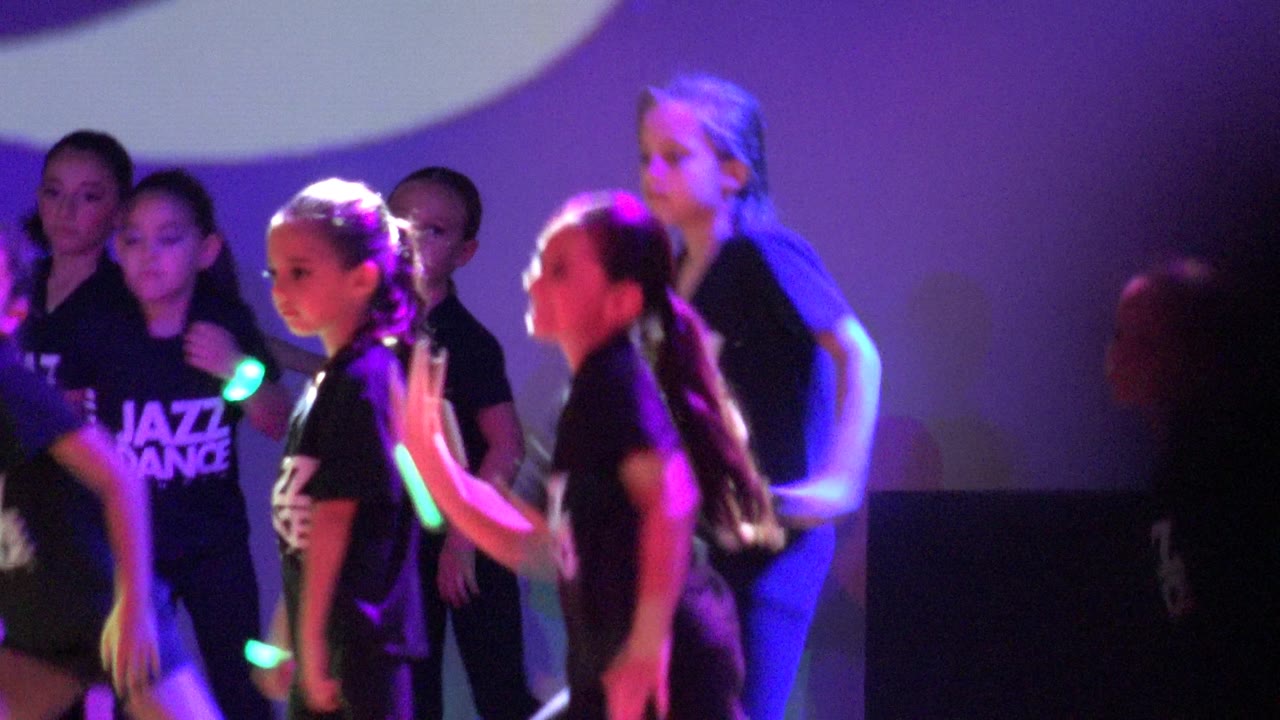 Children in black Jazz Dance shirts stand close together during the Mexico stage routine, with one dancer in profile and purple lighting across the backdrop.