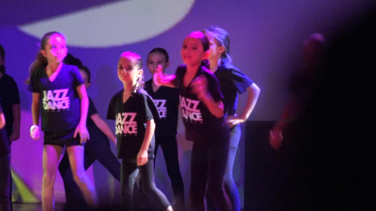 Children in black Jazz Dance shirts begin a stage routine in San Jose Del Cabo BCS Mexico, with purple lighting, a bright backdrop shape, and several visible faces.