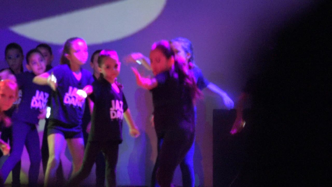 Children in black Jazz Dance shirts dance on a stage in San Jose del Cabo, Mexico, with purple lighting and a dark soft foreground edge at right.