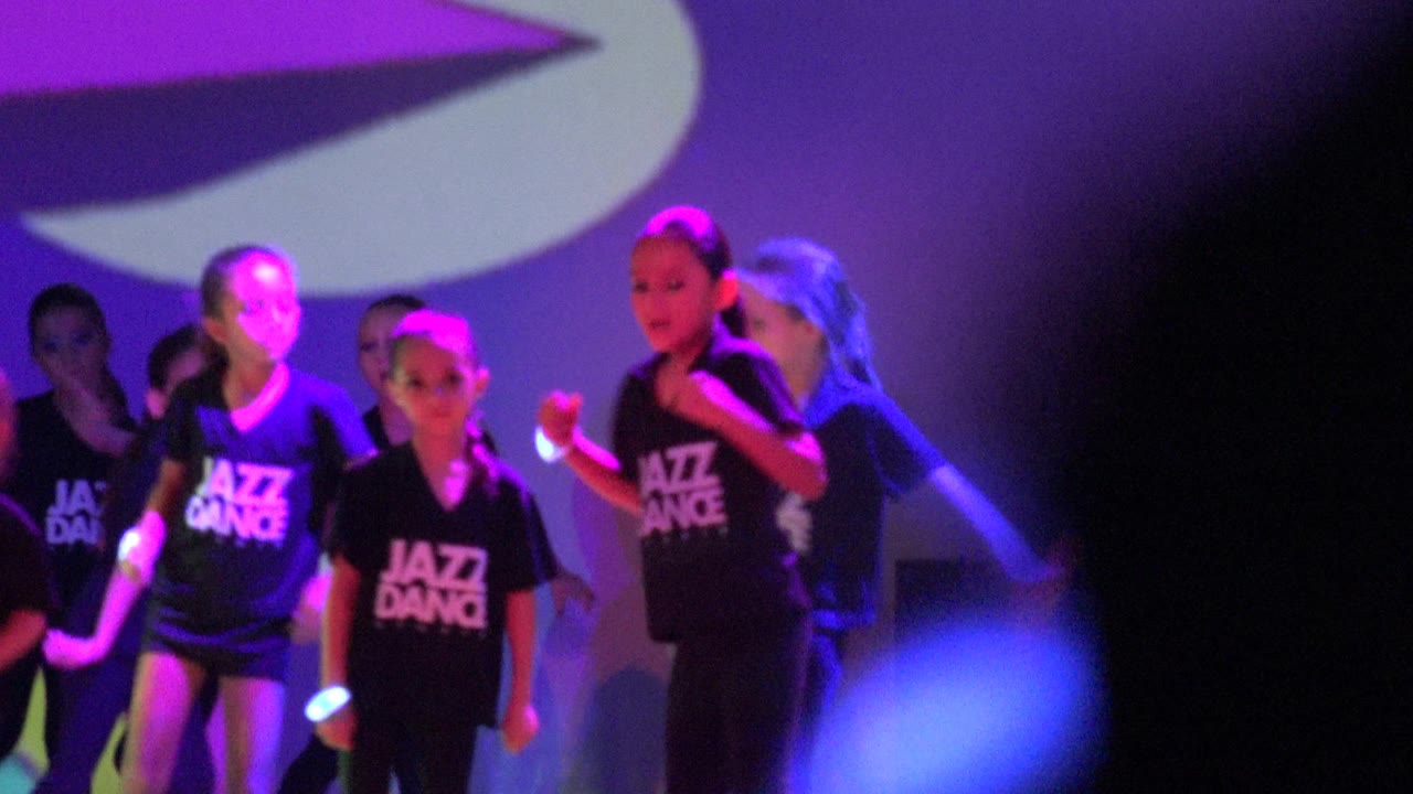 Children continue the jazz dance routine on a stage in San Jose del Cabo, Mexico, with several faces visible and a large dark foreground shape partly blocking the right side.