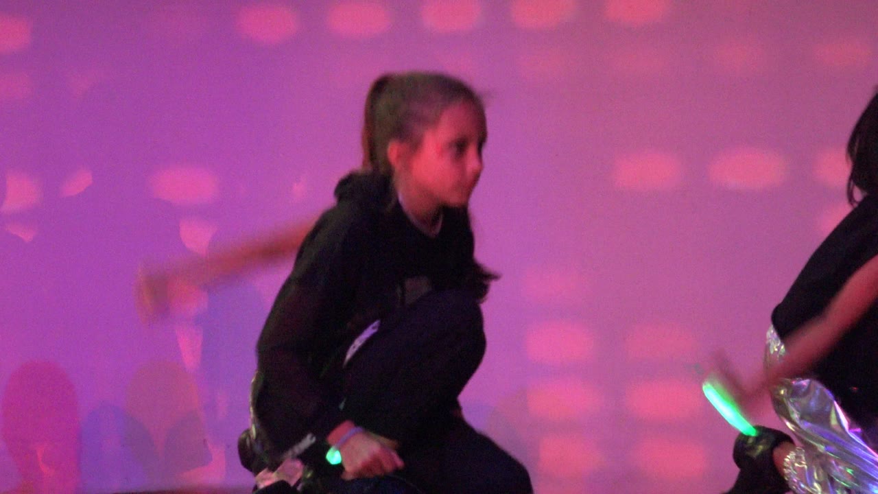 A girl in black stage clothing crouches during a dance performance in San Jose del Cabo BCS Mexico, with pink-purple lighting and another dancer partly visible beside her.