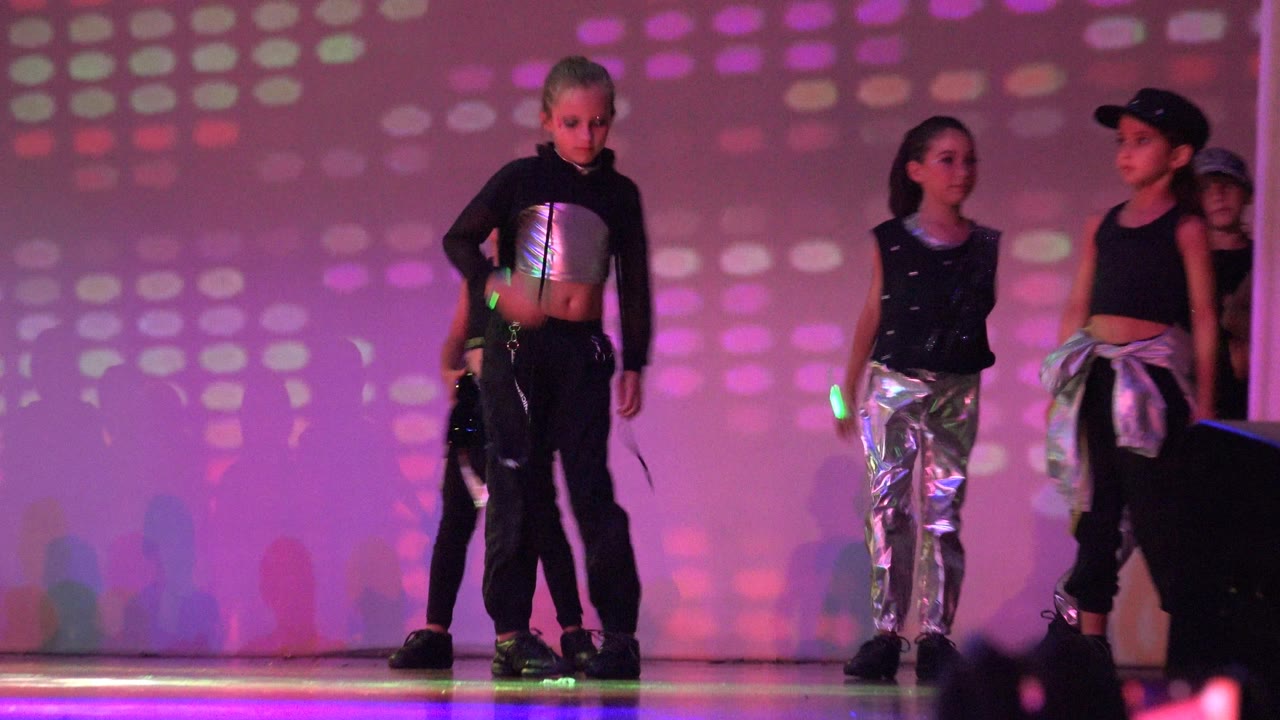 A small group of girls stands on the stage in San Jose del Cabo BCS Mexico, wearing black and silver dance outfits under pink and purple patterned lighting.
