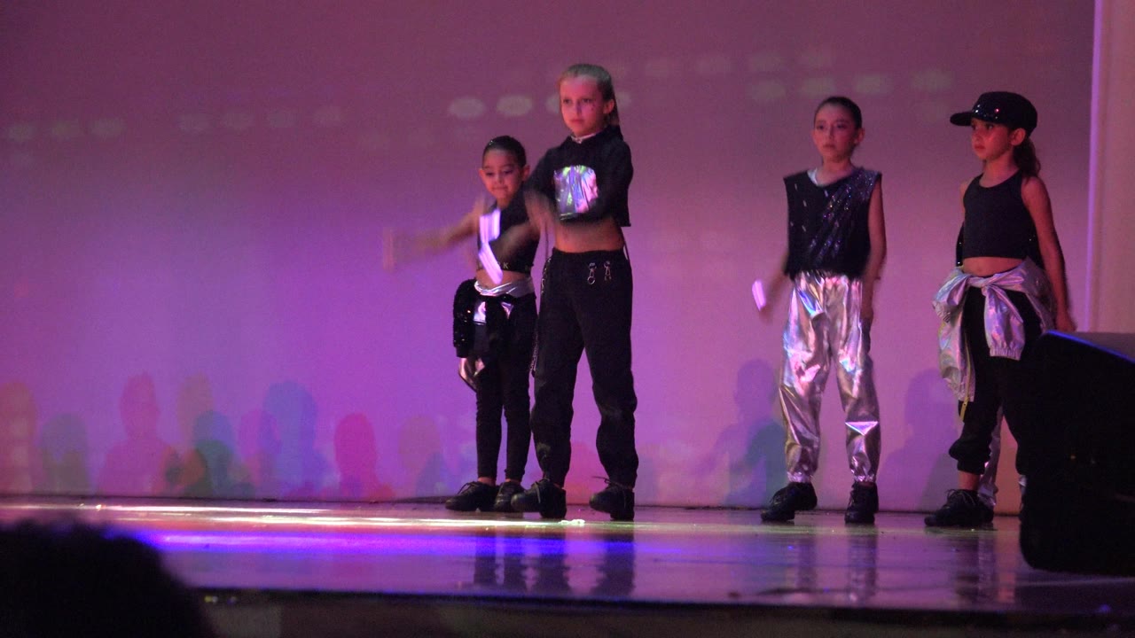 The group continues the San Jose Del Cabo BCS Mexico stage routine, with one child moving at frame left and a small glowing prop visible against the purple-lit background.