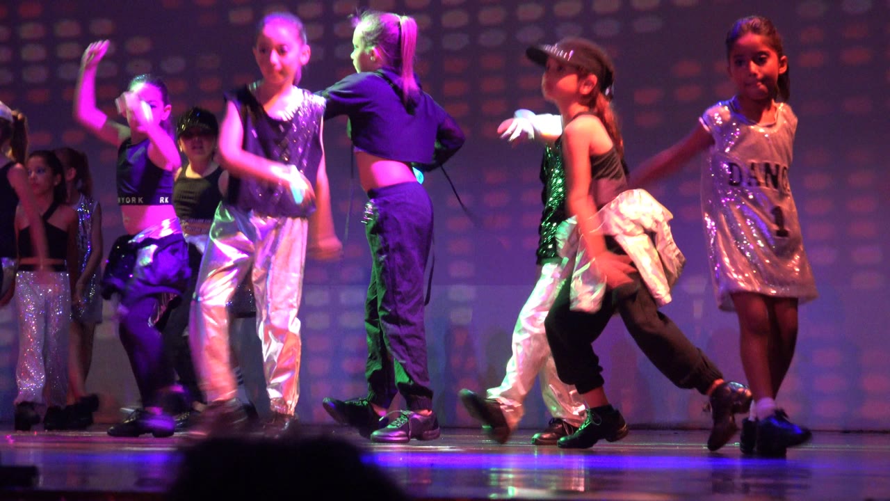 Children perform on a stage in San Jose Del Cabo BCS Mexico, with silver pants, black dancewear, sequins, and purple-green lighting across the backdrop.