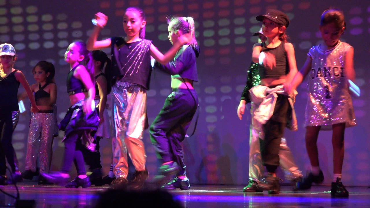 Several children continue the routine on the San Jose Del Cabo BCS Mexico stage, with one dancer in a shiny silver dress near front right and others in black and silver costumes behind.