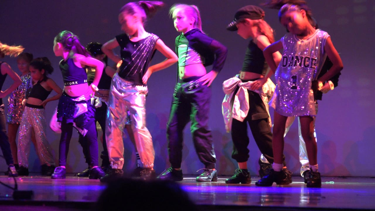 Children stand and turn in a center-stage grouping in San Jose del Cabo BCS Mexico, with green and purple light washing over black, white, and silver dancewear.