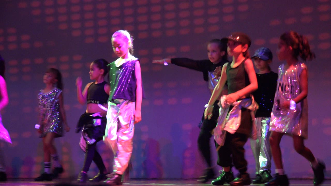 Children in black and silver dance outfits stand and move across a stage in San Jose Del Cabo BCS Mexico with purple and pink patterned lighting behind them.