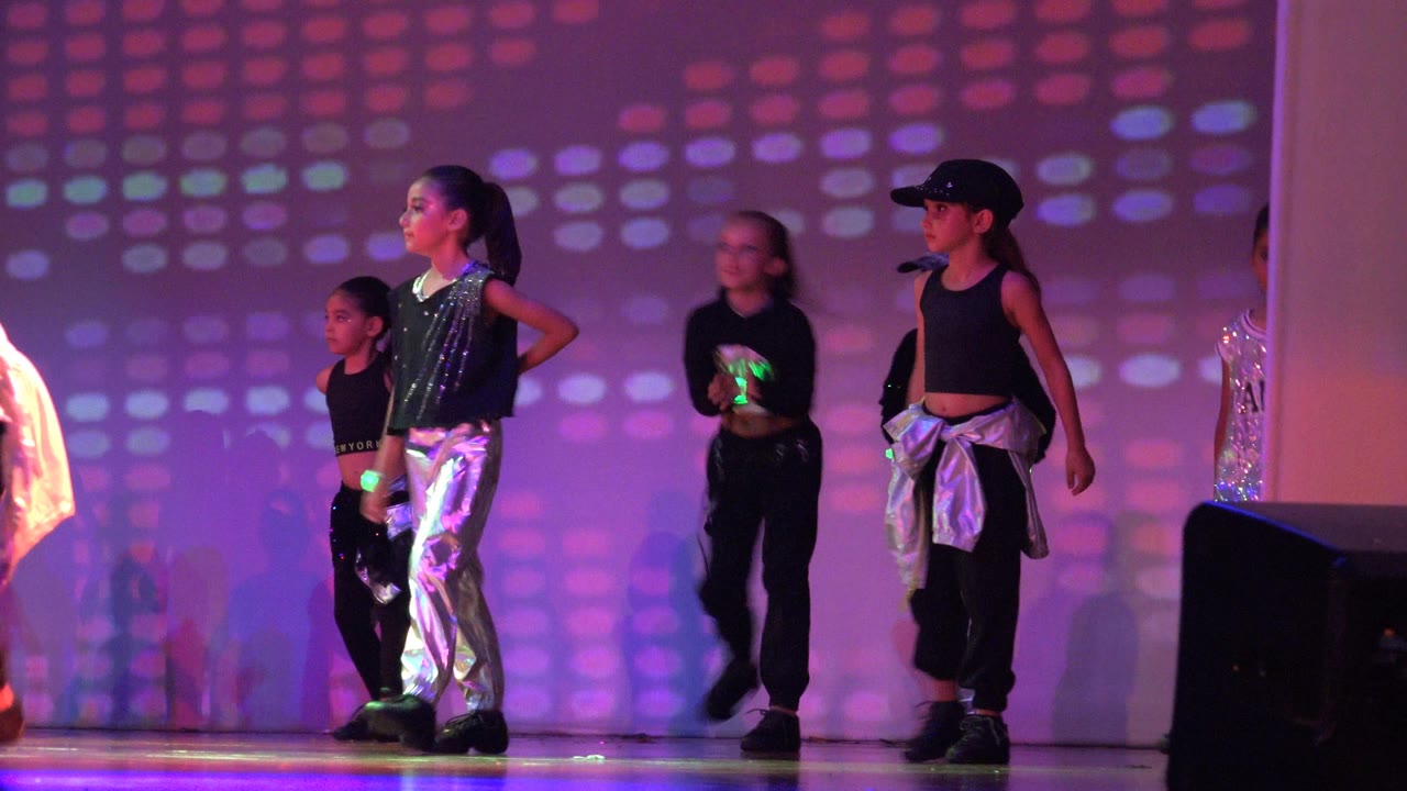 A line of children stands in a stage formation in San Jose Del Cabo BCS Mexico, with black outfits, shiny silver fabric, and magenta dotted light on the wall.