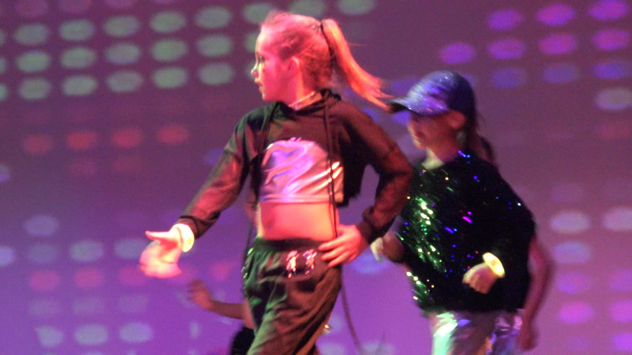 Young dancers perform on a stage in San Jose Del Cabo BCS Mexico, with a blond dancer in the foreground and purple-green light patterns on the back wall.