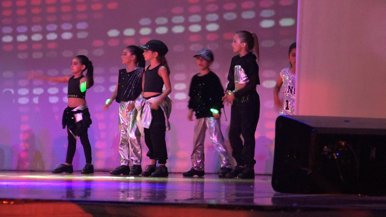 Young dancers in black and metallic costumes stand under pink and purple stage lights in San Jose Del Cabo BCS Mexico, with a speaker blocking part of the lower right frame.