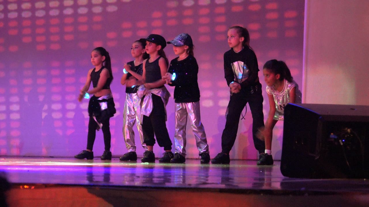 Children stand in formation on a stage in San Jose del Cabo BCS Mexico, with purple lighting, reflective costumes, and a black speaker in the foreground.