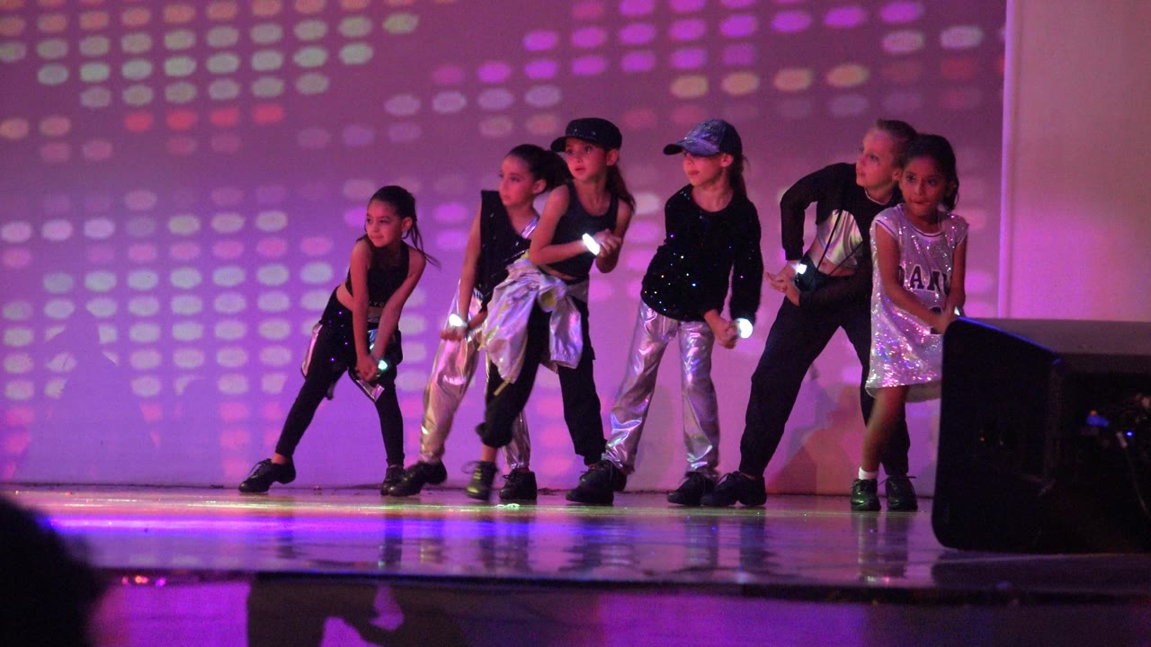 Children in black, silver, and white outfits crouch and gesture on a purple-lit stage in San Jose del Cabo BCS Mexico, with the patterned wall behind them.