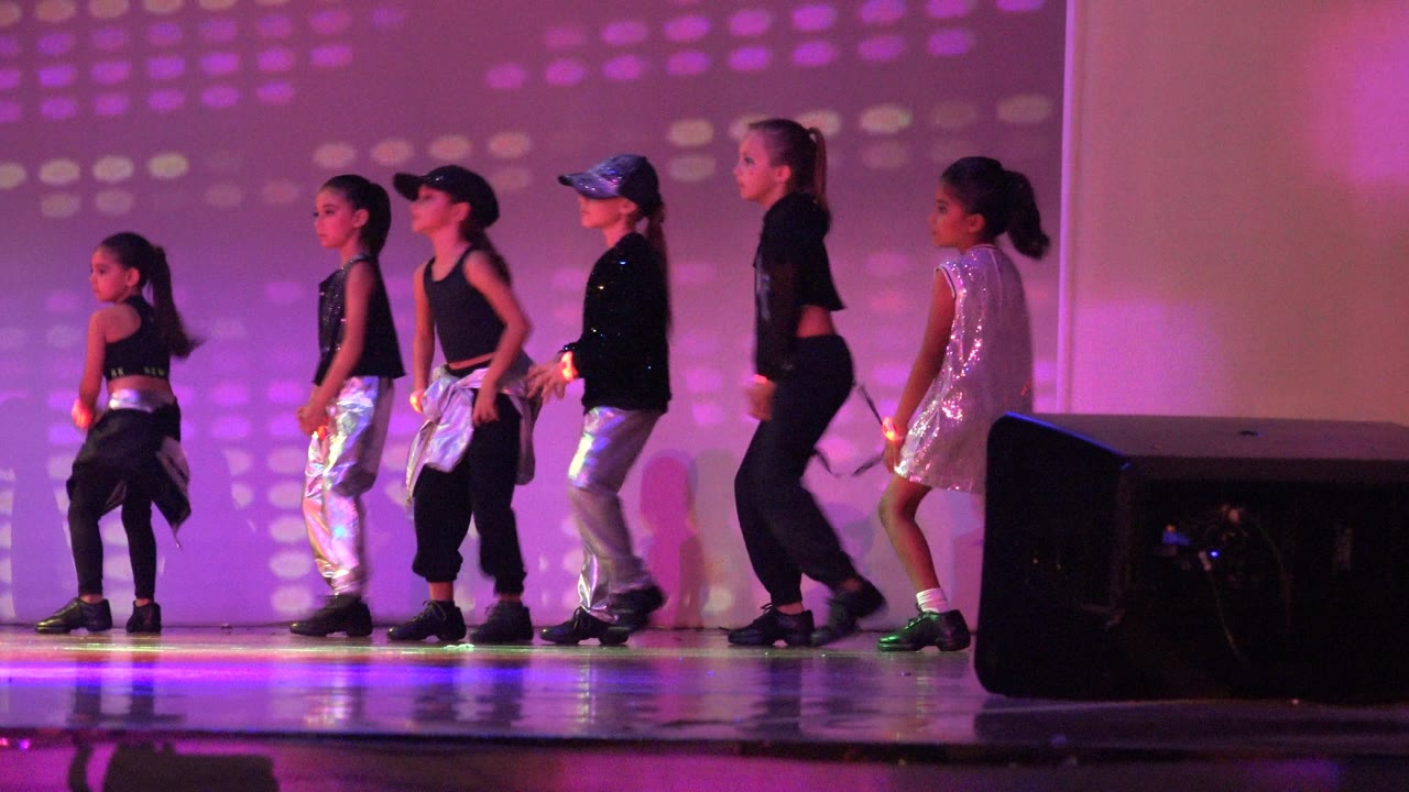 Children in black and silver dance costumes stand in a line on a purple-lit stage in San Jose del Cabo, BCS, Mexico, with a black speaker visible near the front edge.