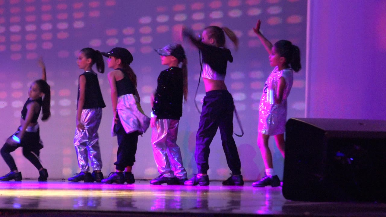 Children move across the stage with arms raised under purple lights.