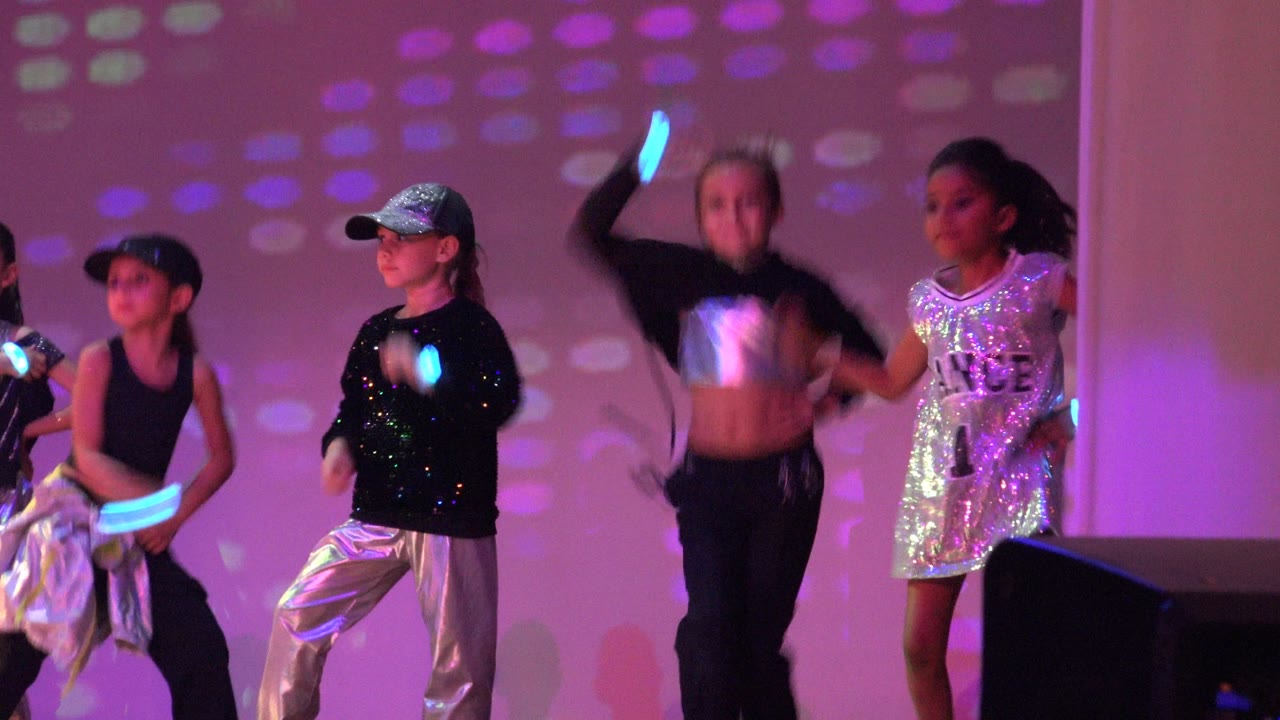 Children perform on a stage in San Jose Del Cabo BCS Mexico, with one child in a silver cap and pants, another in black, and purple-pink projected lights behind them.
