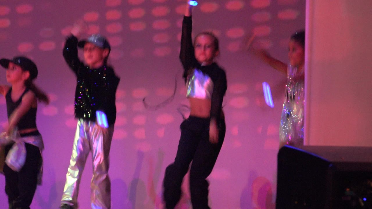 The San Jose Del Cabo BCS Mexico performance continues with children in black and silver costumes dancing near a pale column, a dark stage monitor, and pink-purple projected lights.