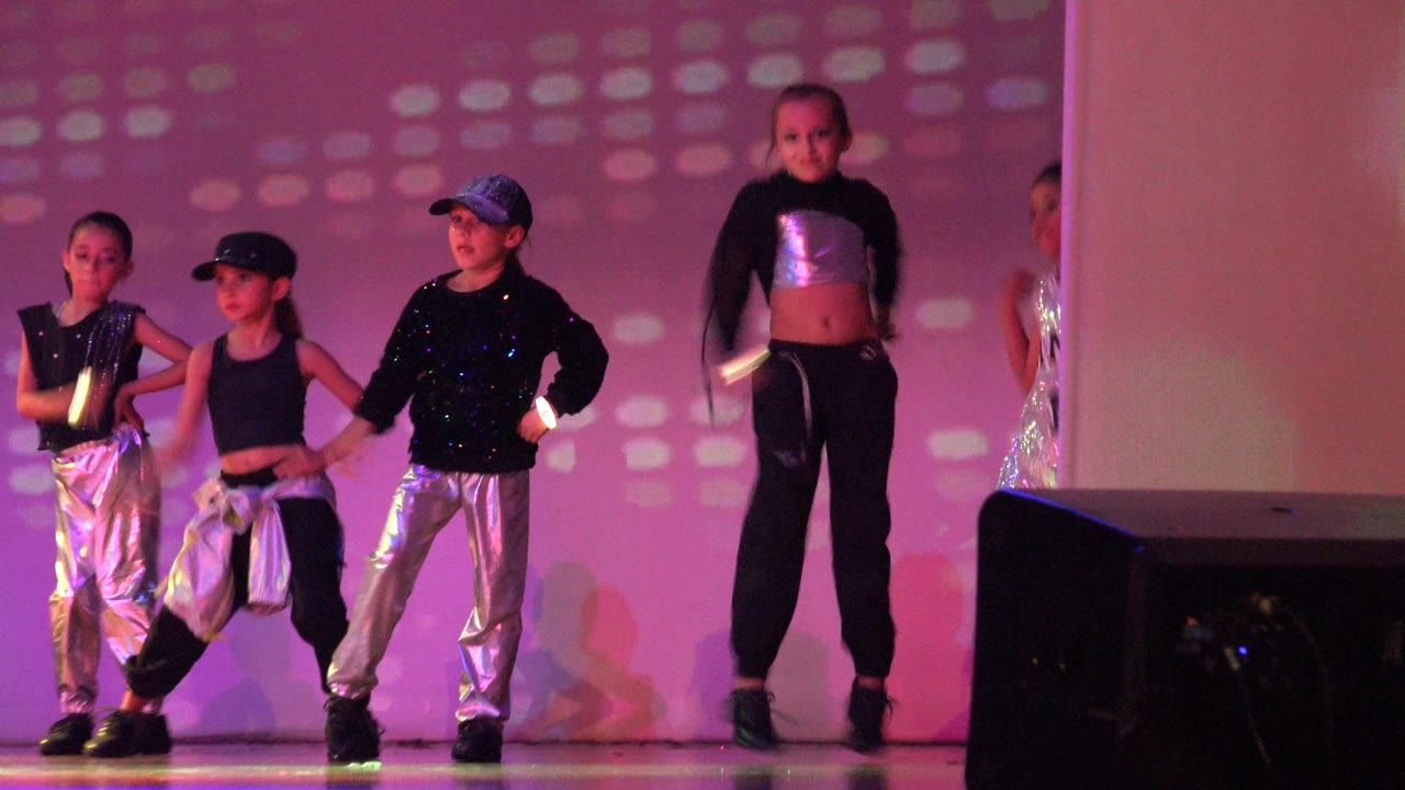 Children perform a synchronized dance routine on a stage in Mexico, with silver pants, black tops, glow sticks, pink lighting, and a foreground speaker visible.
