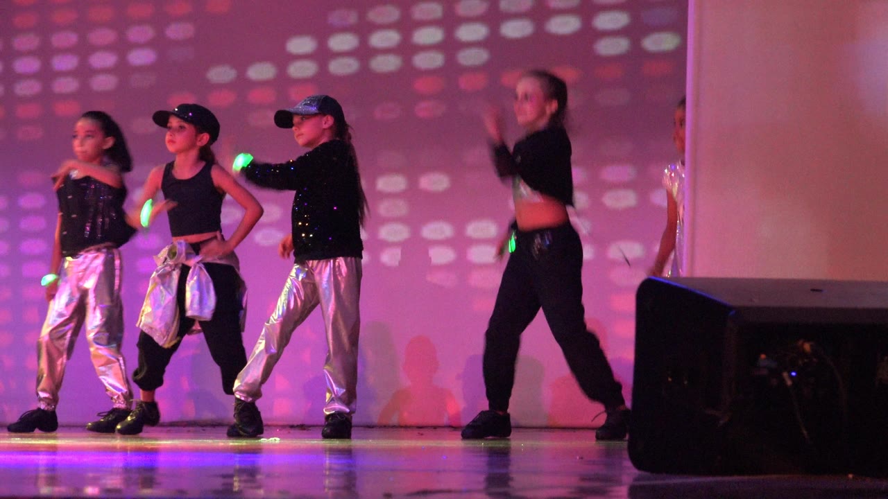 Young dancers on the Mexico stage step through the routine, with one performer extending a glowing stick and others in silver and black costumes under pink light.