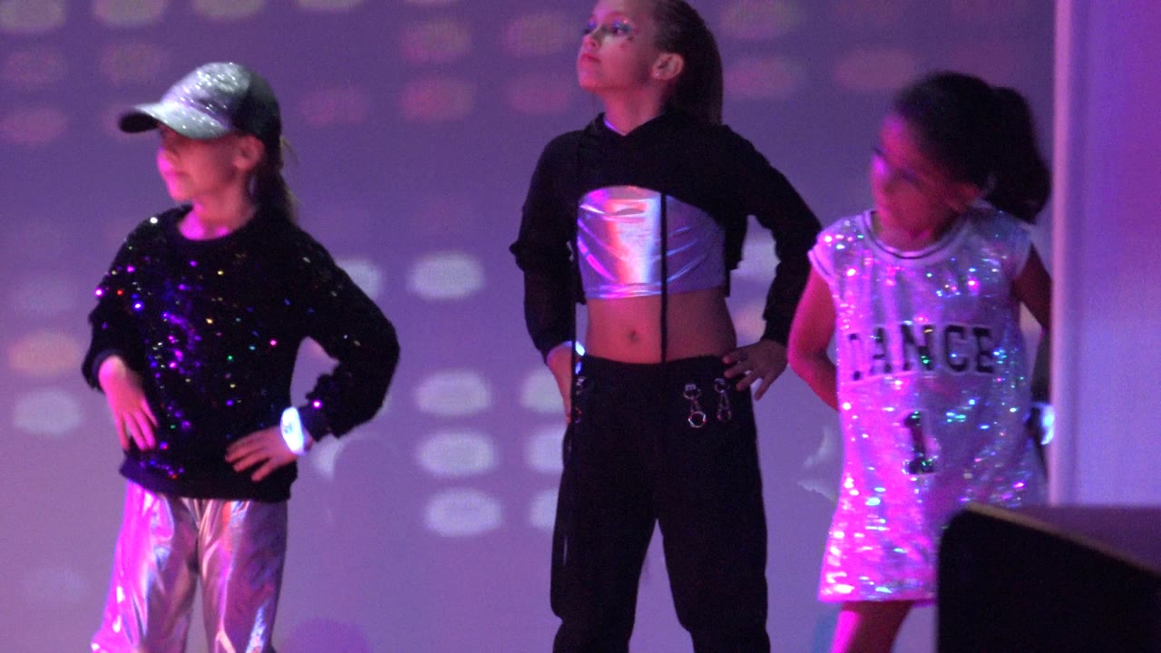 Three children pose on stage in silver and black dance clothing under colored lights.