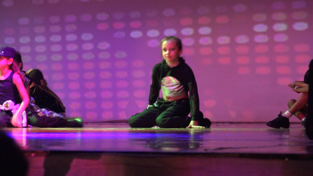 A child dancer sits near center stage in San Jose Del Cabo BCS Mexico, with other children partially visible and a dark stage speaker in the foreground.