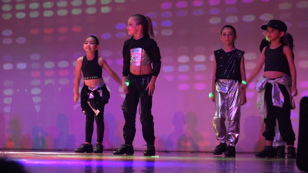 Four children in black and silver dance outfits stand on a lit stage in San Jose Del Cabo BCS Mexico, with glow sticks and patterned purple light behind them.