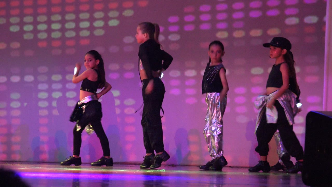 Children hold staged dance poses in San Jose del Cabo BCS Mexico, with silver pants catching purple light and a dark speaker partly visible at the right edge.