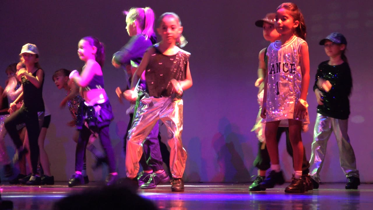 Children continue the dance routine in San Jose Del Cabo BCS Mexico, with one sequined dress near the front and other dancers crossing behind in black and silver outfits.