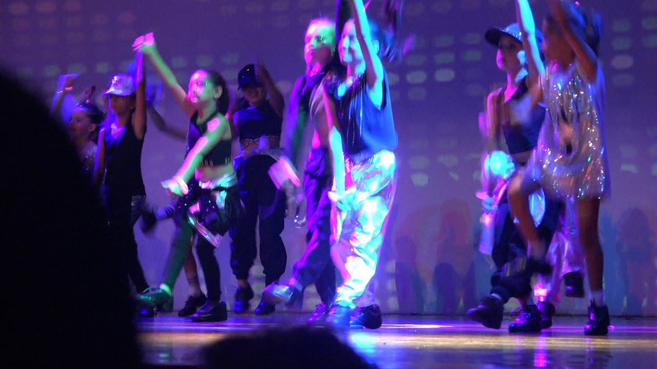 Group of young dancers crossing the stage in shiny outfits under blue and purple light.