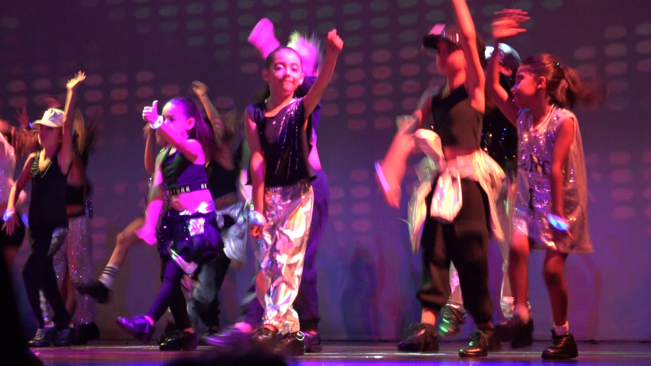 Children dance in San Jose Del Cabo BCS Mexico with multiple arms lifted, shiny outfits catching pink and blue light, and the patterned stage backdrop behind them.
