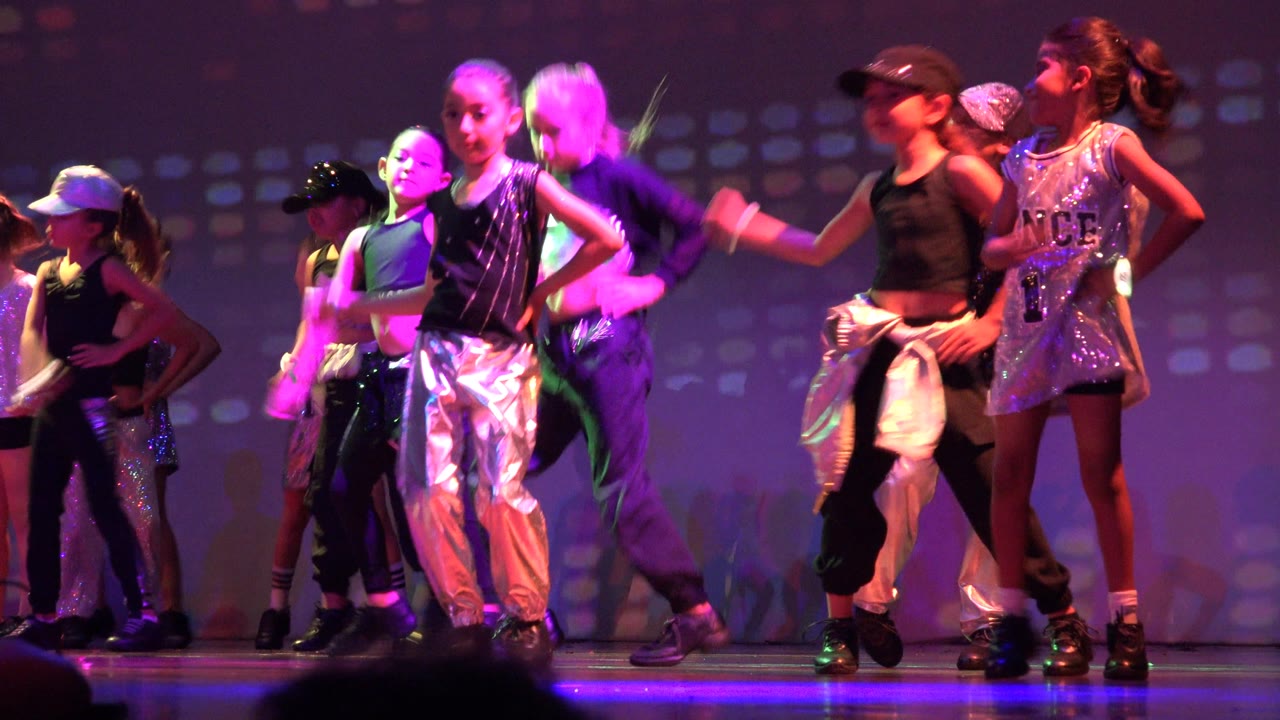 Children perform a synchronized stage routine in San Jose Del Cabo BCS Mexico, with silver pants, black tops, sequined costumes, and purple-pink light on the backdrop.