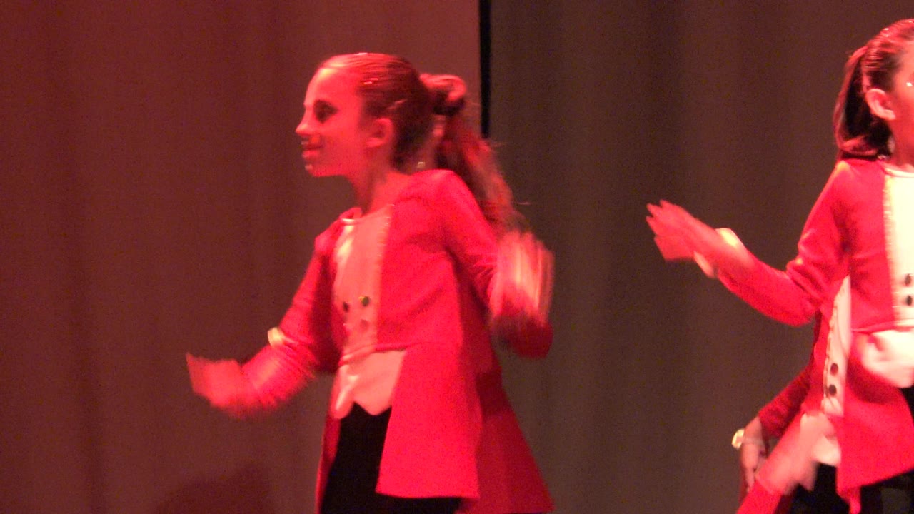 A girl in a red jacket and white top faces left during the stage routine in San Jose Del Cabo BCS Mexico, with glowing wrist lights and another dancer behind her.