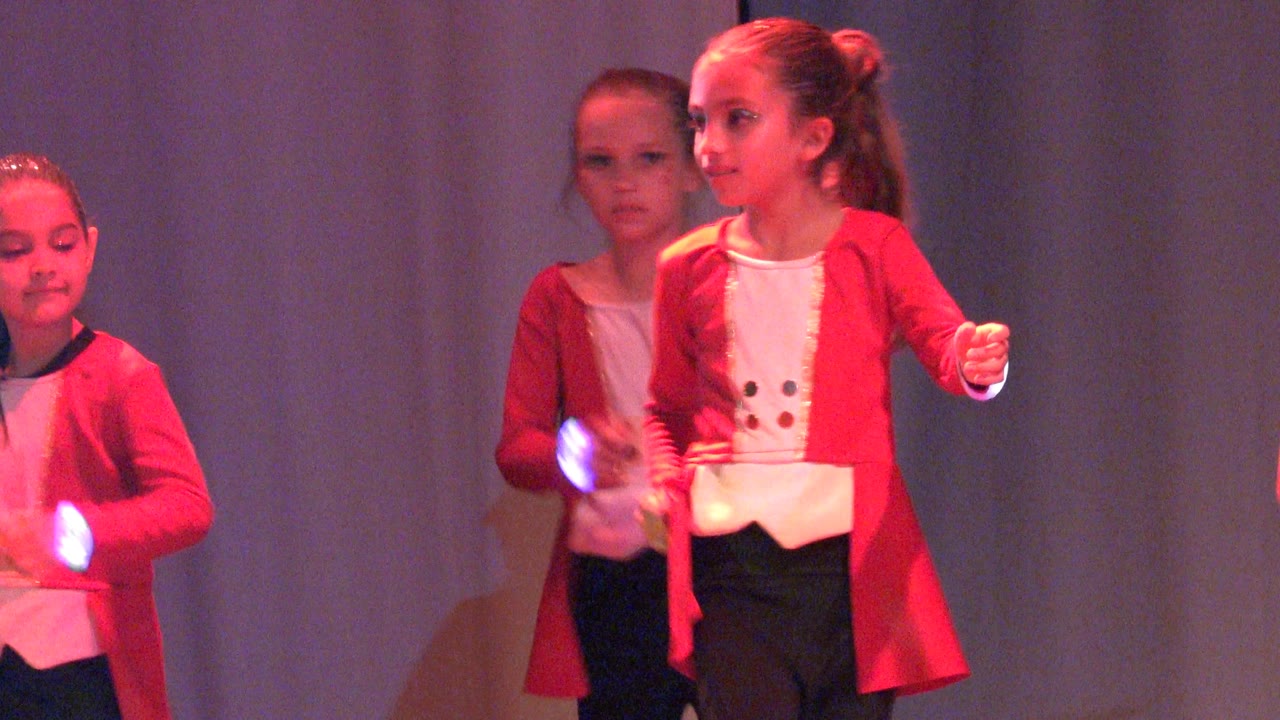 Children in red jackets and white tops begin a stage dance moment in San Jose Del Cabo BCS Mexico, with faces visible and red lighting on a dark curtain backdrop.