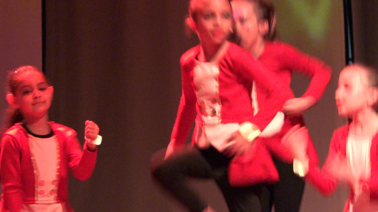 Two children in matching red jackets and white tops hold positions during the dance in San Jose Del Cabo BCS Mexico, with warm red stage light across their faces and costumes.