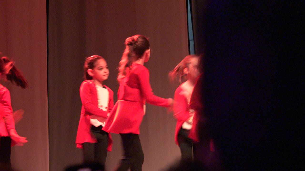 Children in red tops and black pants perform on a stage in Mexico, with a curtain backdrop and red stage lighting at the start of the clip.