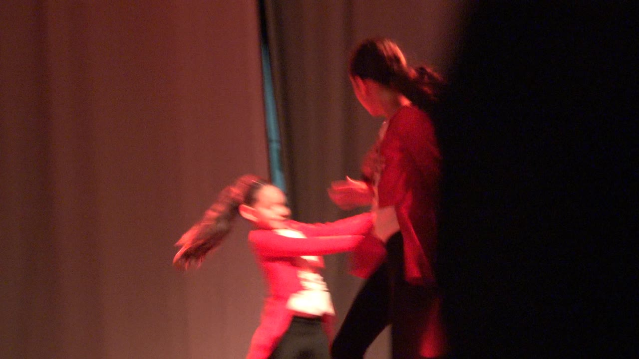 A young dancer in red reaches toward a taller performer at the right edge of the stage, with the curtain background lit red in Mexico.