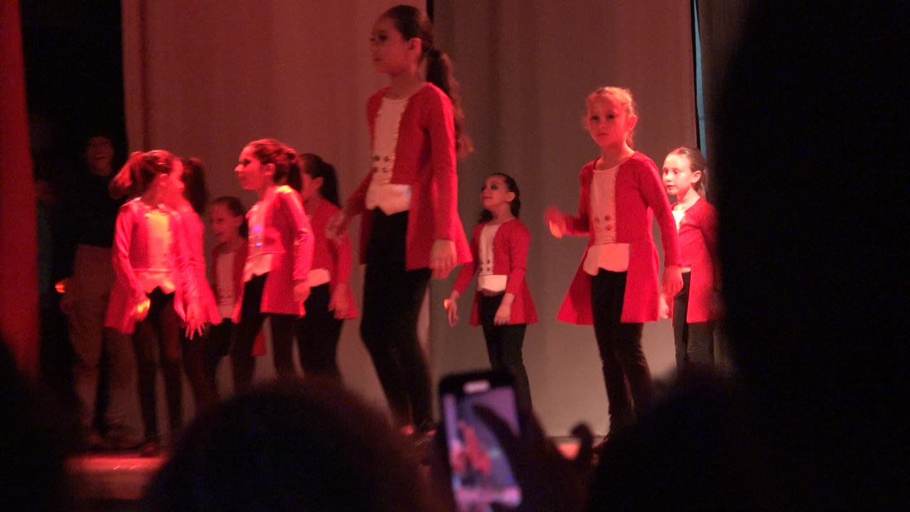 Girls in red jackets and black pants perform on a stage in Mexico as audience silhouettes and a phone screen appear in the foreground.