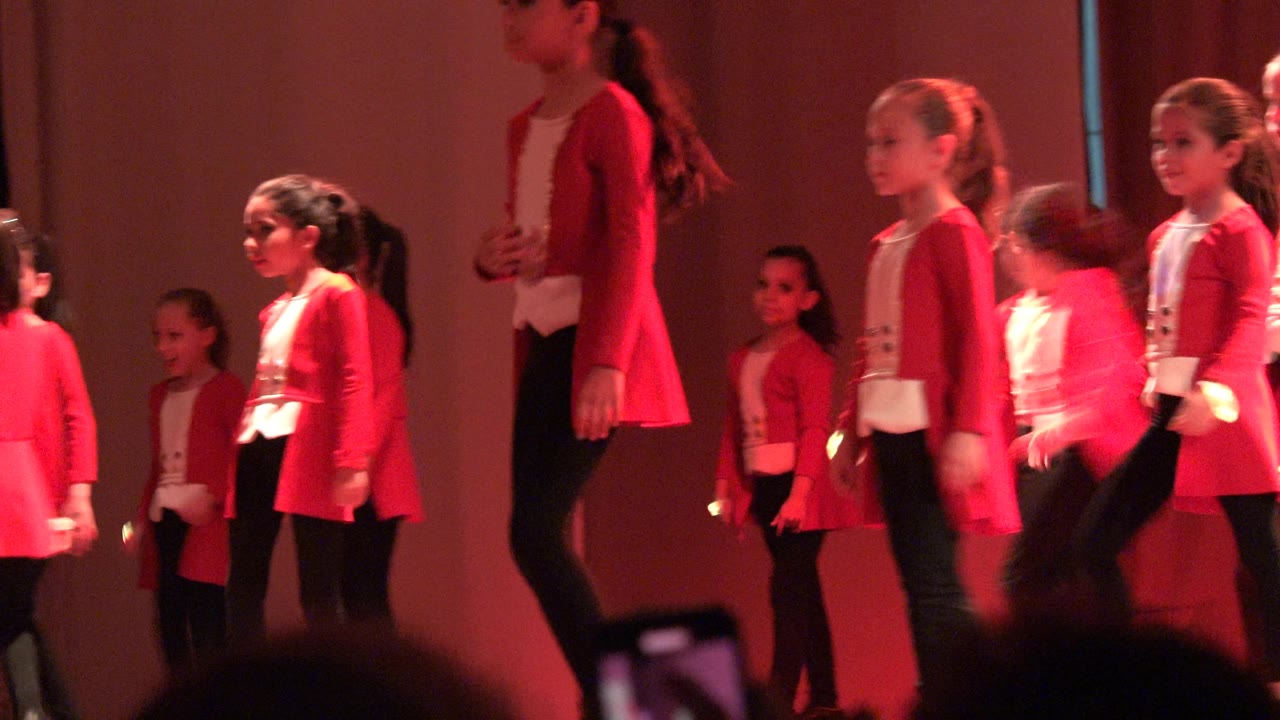 Several girls in matching red jackets and black pants shift positions on the Mexico stage while audience silhouettes remain along the bottom edge.