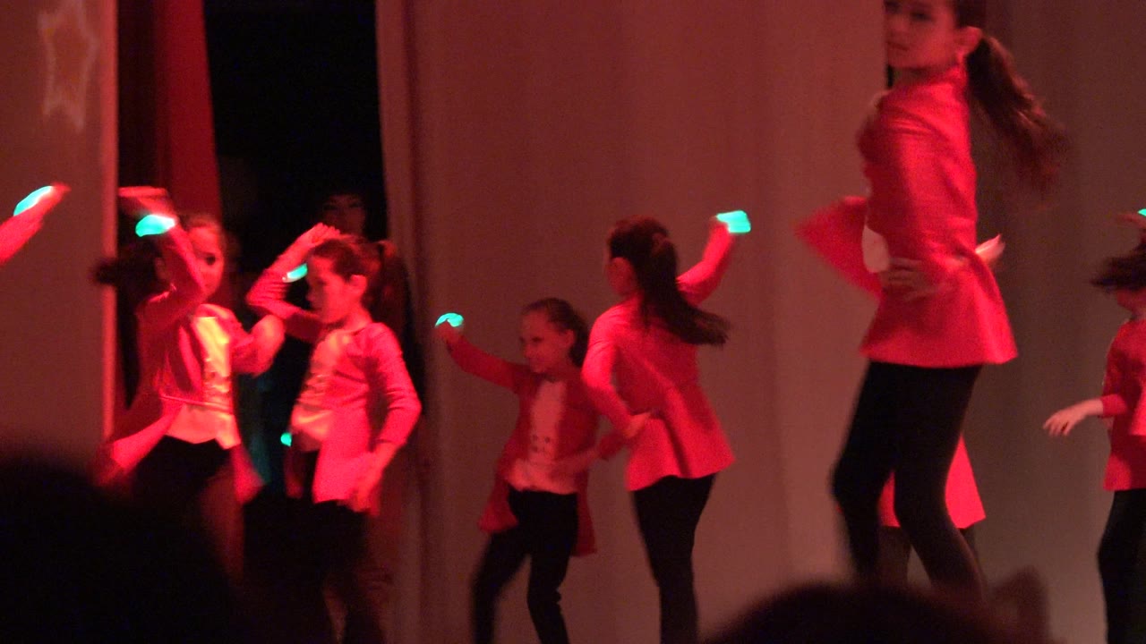 Children perform on a red-lit stage in San Jose del Cabo, Mexico, with red jackets, black pants, pale curtains, and audience silhouettes along the bottom edge.