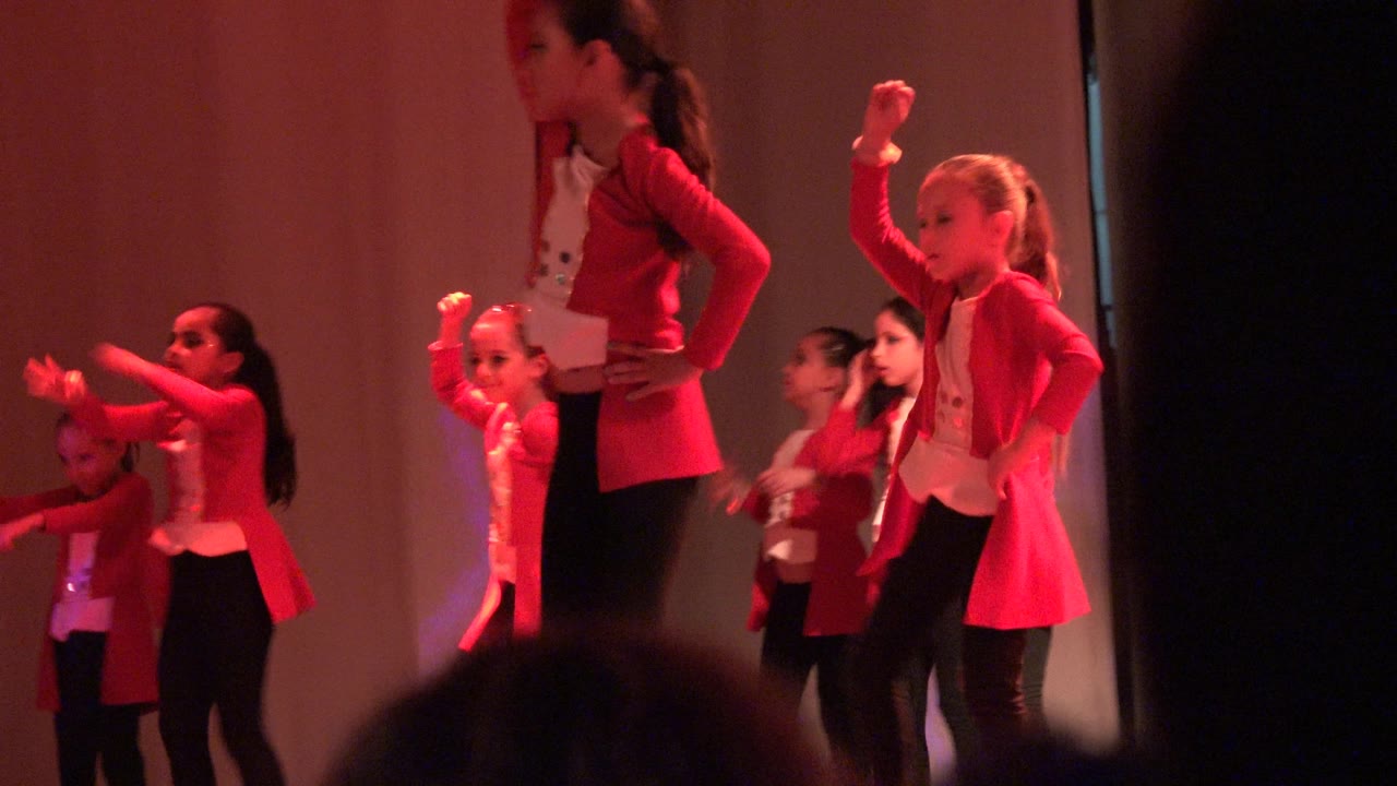 Children perform a staged routine in San Jose del Cabo, Mexico, with red costumes, black pants, glowing handheld props, and dark audience heads cutting into the lower frame.