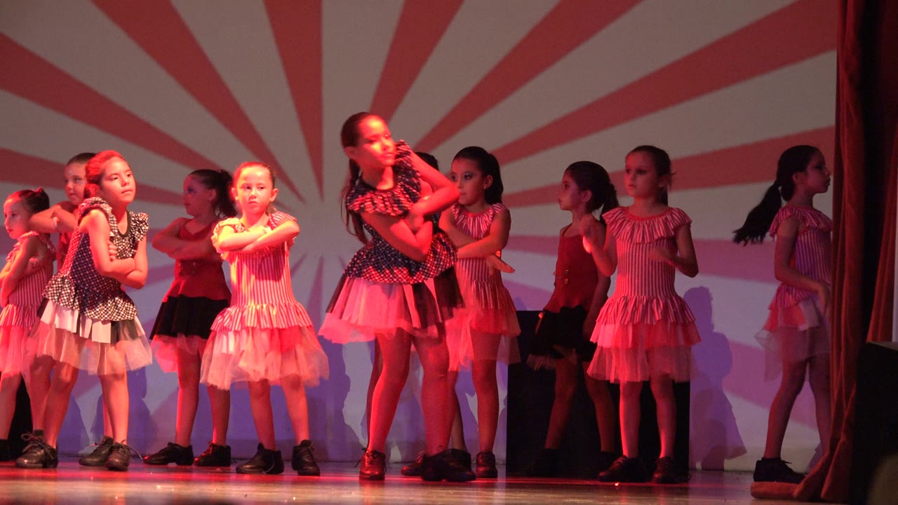 Young dancers in San Jose del Cabo BCS Mexico stand across a stage in pink, red, black, and white outfits as red light washes over a striped backdrop.