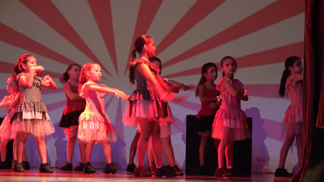 A girl in a black and white dress stands near the front while other young dancers in pink and red costumes continue the routine in San Jose del Cabo BCS Mexico.